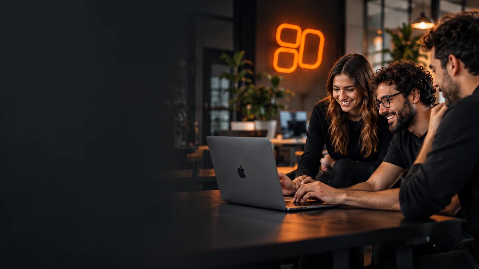 Three WEBSTYLE team members smiling at a laptop in a dark studio with an orange neon logo on the wall